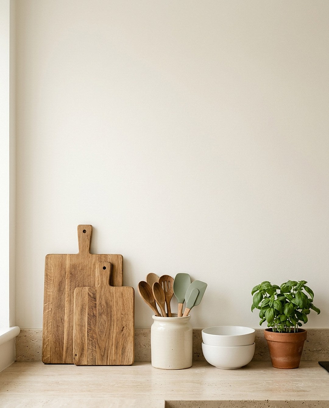 Wooden cutting boards, a crock of utensils, white bowls and a basil plant on a kitchen counter